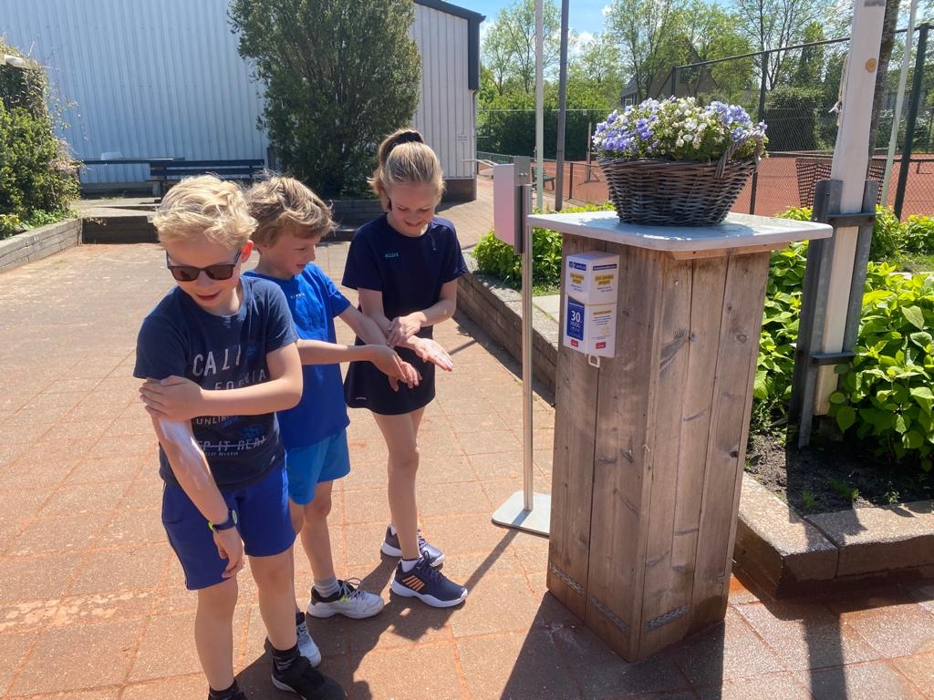 drie kinderen bij zonnebrandcreme dispenser en tennisbaan kinderen smeren zich in met carelyn zonnebrandcreme